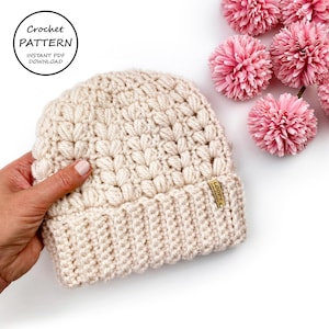 May include: Cream-coloured crochet beanie with a textured stitch pattern. The hat has a folded brim and a small tag. The image includes the text "Crochet Pattern" and "Instant PDF Download" in a circle. Pink pom-pom flowers are in the background.