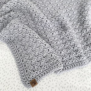 May include: A soft, gray crocheted blanket with a textured, puffy design. The blanket is folded and has a small brown tag with the word "Love" on it.