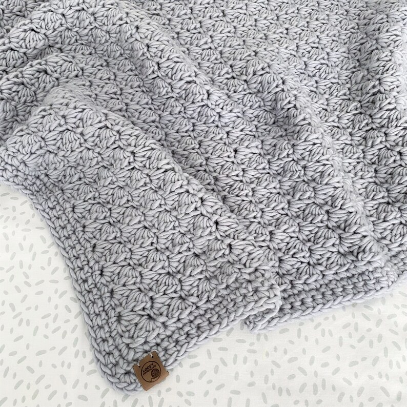 May include: A soft, gray crocheted blanket with a textured, puffy design. The blanket is folded and has a small brown tag with the word "Love" on it.
