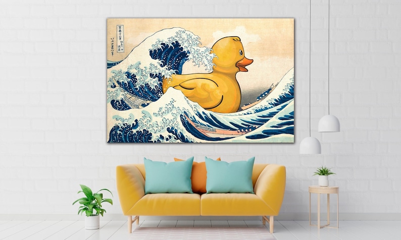 May include: A canvas print depicting a yellow rubber duck in a traditional Japanese wave scene. The duck floats on the crest of a large blue wave, with a light beige sky in the background. The artwork is displayed above a yellow sofa with teal pillows.