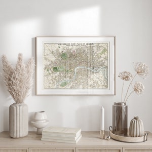 May include: A framed vintage map of London titled "Whitbread's New Plan of London" with a white background and black text. The map is hanging on a white wall above a wooden shelf with a vase of dried pampas grass, a stack of books, a candle, a vase of dried flowers, and a decorative tray.