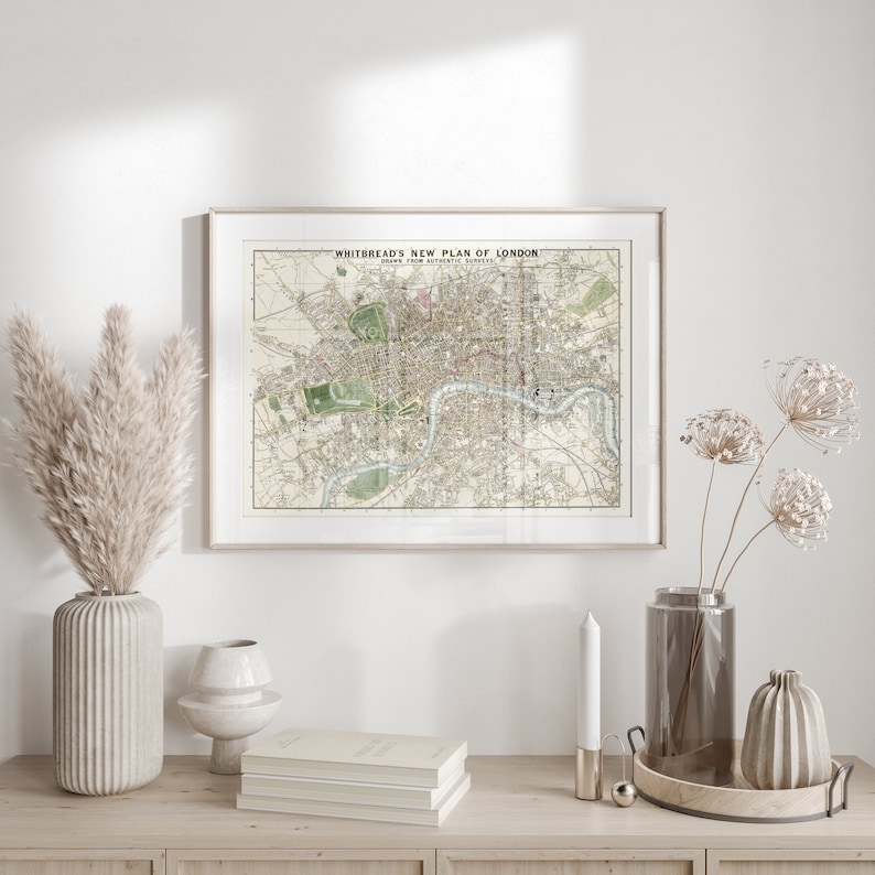 May include: A framed vintage map of London titled "Whitbread's New Plan of London" with a white background and black text. The map is hanging on a white wall above a wooden shelf with a vase of dried pampas grass, a stack of books, a candle, a vase of dried flowers, and a decorative tray.