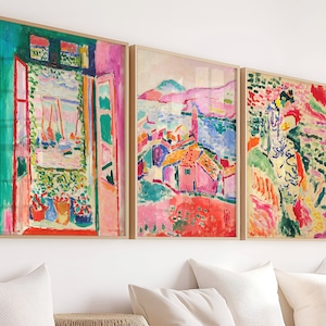 Op de afbeelding: Drie ingelijste kunstprints in een kleurrijke, abstracte stijl. De eerste print toont een raam met uitzicht op een haven en boten. De tweede print toont een dorp met huizen en een waterpartij. De derde print toont een vrouw in een kleurrijke jurk die in een landschap staat.
