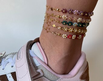 May include: A close-up of a person's ankle wearing multiple beaded anklets. The anklets are made with small gold stars and colorful flower-shaped beads. The person is wearing a pair of pink and brown Adidas sneakers.
