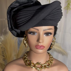 Puede incluir: Un turbante negro con pliegues horizontales estructurados y un gran detalle fruncido en el lateral. El turbante se combina con un collar de eslabones dorados gruesos y pendientes de eslabones a juego sobre un busto de maniquí.
