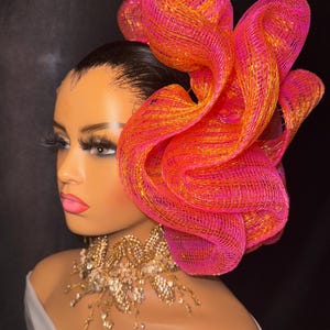Puede incluir: Un accesorio para el cabello de malla rosa y naranja con un gran diseño rizado. El accesorio se lleva en un maniquí con una blusa blanca y un collar de cuentas doradas.