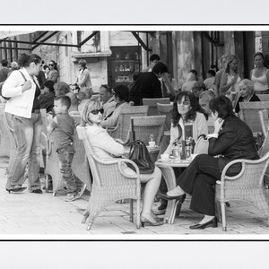 Könnte beinhalten: Ein Schwarzweißfoto von Menschen, die an Tischen im Freien sitzen. Die Tische sind aus Holz und haben Korbstühle. Die Leute tragen Freizeitkleidung und einige tragen Sonnenbrillen. Im Hintergrund sind Pflanzen und Bäume zu sehen.