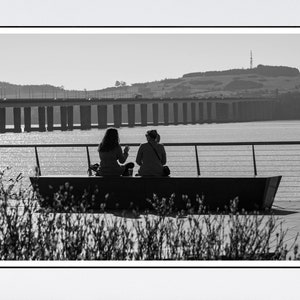 May include: Two people sit on a bench overlooking a body of water with a bridge in the distance. The bridge has many pillars and is made of concrete. The people are silhouetted against the water and the bridge.