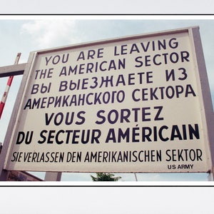 Könnte beinhalten: Ein weißes Schild mit schwarzem Text in Englisch, Russisch, Französisch und Deutsch, das "Sie verlassen den amerikanischen Sektor" lautet. Das Schild ist an einem Pfosten befestigt und trägt unten das Logo der US-Armee.