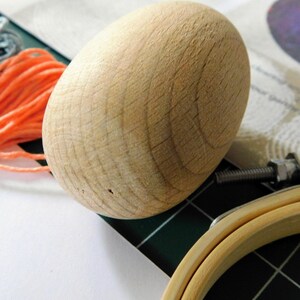 May include: A close-up of a wooden egg-shaped object, with a natural wood grain pattern. The egg is resting on a green surface, with a wooden embroidery hoop in the background.