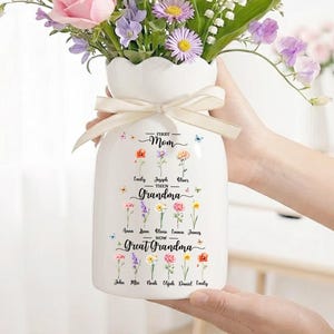 May include: A white ceramic vase with a scalloped rim, filled with a vibrant bouquet of flowers. The vase is adorned with a cream-colored ribbon and features text that reads "First Mom Then Grandma Now Great Grandma."