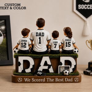 May include: A soccer-themed figurine featuring a father and three children, all wearing matching jerseys. The word "DAD" is prominently displayed with soccer ball accents. The base reads "We Scored The Best Dad."