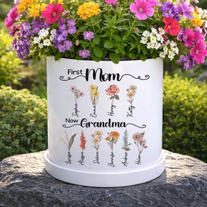May include: A white ceramic flower pot filled with vibrant, colorful flowers. The pot is inscribed with the words "First Mom Now Grandma" and features floral illustrations with names. The pot sits on a white saucer.