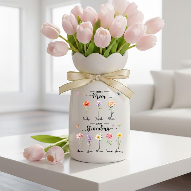 May include: A white ceramic vase with a cream-colored ribbon holds a bouquet of light pink tulips. The vase is decorated with floral illustrations and the text "First Mom Now Grandma" with names below.