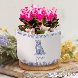 May include: A white ceramic planter with a blue floral design and a seated dog illustration, labeled "Birdie." The planter holds a succulent with vibrant pink flowers. It sits on a wooden coaster.