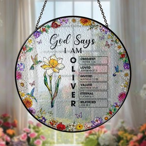 May include: Round stained glass window art with a black metal frame and hanging chain. The design features the text "God Says I AM OLIVER" with biblical references. A daffodil, hummingbirds, butterflies, and floral border complete the design.