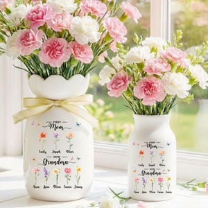 May include: Two white ceramic vases filled with pink and white carnations. The larger vase has a scalloped rim and a yellow ribbon. Text on the vases reads "First Mom Now Grandma" with names and flower illustrations.