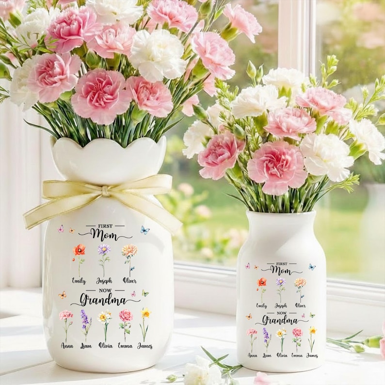 May include: Two white ceramic vases filled with pink and white carnations. The larger vase has a scalloped rim and a yellow ribbon. Text on the vases reads "First Mom Now Grandma" with names and flower illustrations.