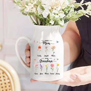 May include: White ceramic pitcher with a handle, decorated with colorful floral illustrations and the words "First Mom - Now Grandma." The pitcher is filled with white flowers. Butterflies are also featured in the design.