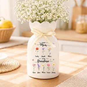 May include: A white ceramic vase with a scalloped rim, tied with a cream-colored ribbon, filled with white flowers. The vase is decorated with floral illustrations and the text "First Mom Now Grandma" with names below.