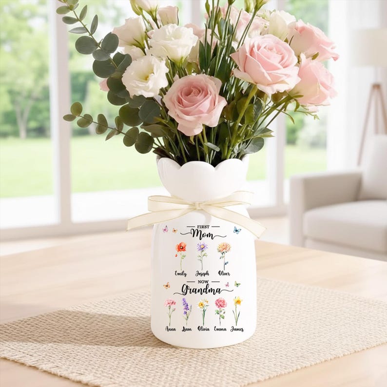 May include: White ceramic vase with a scalloped rim, tied with a cream ribbon, and filled with pink and white flowers. The vase is decorated with floral illustrations and the text "First Mom - Now Grandma" with names below.
