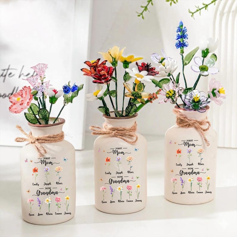 May include: Three cream-colored vases with colorful glass flowers. Each vase has a jute twine bow and the text "First Mom Now Grandma" with names and flower illustrations. The vases hold various flowers.
