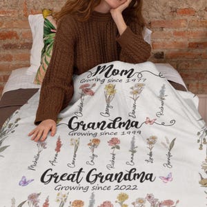 May include: White floral blanket with the text "Mom Growing since 1977", "Grandma Growing since 1999", and "Great Grandma Growing since 2022". The blanket features a floral design with names written on each flower.