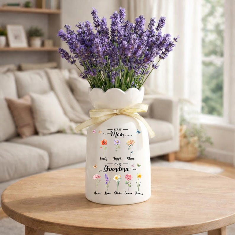 May include: A white ceramic vase with a scalloped rim, tied with a cream-colored ribbon, holds a bouquet of purple lavender. The vase is decorated with floral illustrations and the text "First Mom Now Grandma".