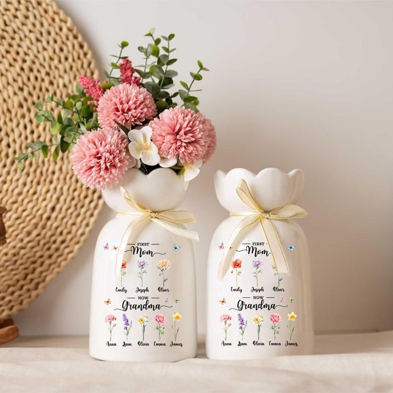 May include: Two white ceramic vases with a floral design and the text "First Mom Now Grandma". One vase holds pink flowers and greenery. Each vase has a gold ribbon tied around the neck. The vases are decorated with colorful flowers and butterflies.