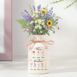 May include: A cream-colored vase with a bouquet of colorful flowers, including sunflowers, daisies, and lavender. The vase is tied with twine and has the text "First Mom - Now Grandma" with names of family members. A thoughtful gift.