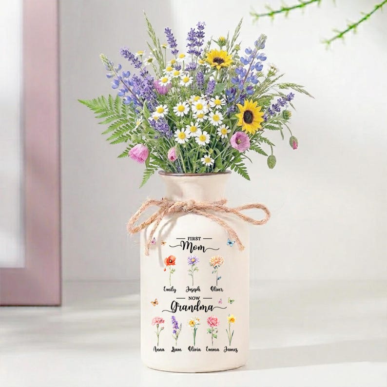 May include: A cream-colored vase with a bouquet of colorful flowers, including sunflowers, daisies, and lavender. The vase is tied with twine and has the text "First Mom - Now Grandma" with names of family members. A thoughtful gift.