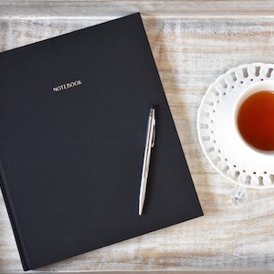 May include: A black notebook with the word "NOTEBOOK" embossed in gold on the cover, a silver pen, and a cup of tea on a white saucer with a decorative lace pattern, all on a wooden tray.