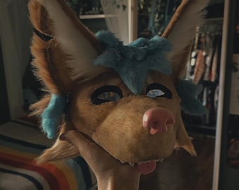 May include: A brown and tan furry fox-like mask with blue accents. The mask has large, pointed ears, black eye openings, and a pink nose. The tongue is visible. The mask is held up by a hand.