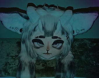 May include: A white and gray furry mask with large ears, horns, and a stylized face. The mask has a painted face with dark eyes, a small nose, and a closed mouth. The mask has long, furry appendages that hang down.