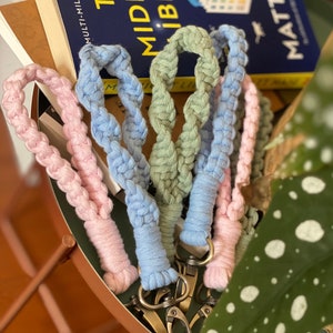 May include: Four braided keychains in pink, blue, green, and blue with silver key rings. The keychains are on a brown surface with a green plant in the background.
