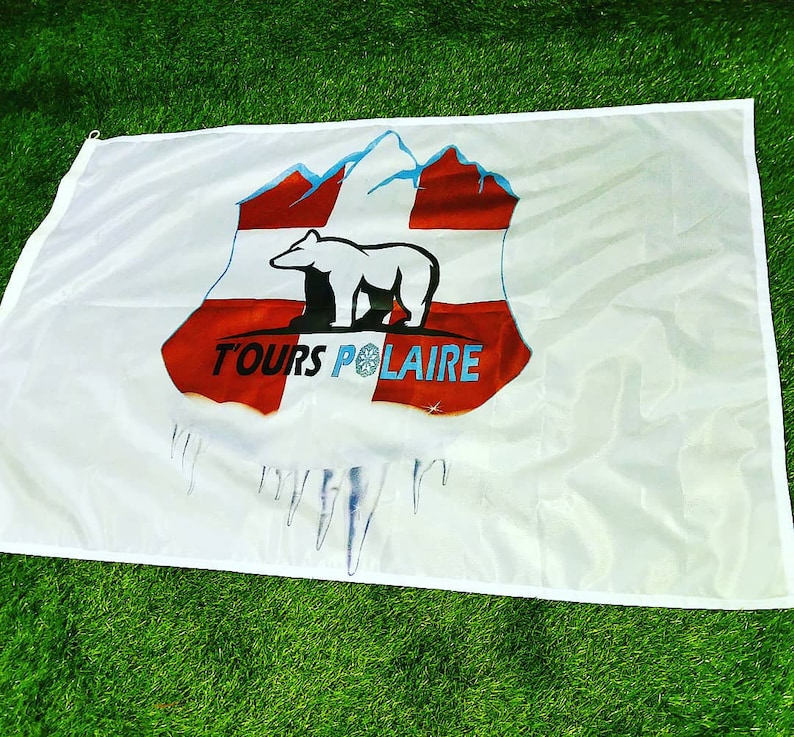 Puede incluir: Bandera blanca con un dise&ntilde;o que presenta un oso polar, monta&ntilde;as y el texto "TOURS POLAIRE" en azul. El dise&ntilde;o incluye elementos rojos y blancos, con car&aacute;mbanos en la parte inferior. La bandera se muestra sobre una superficie verde.
