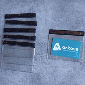 Op de afbeelding: Transparante plastic badgehouders met zwarte bovenkanten. Eén houder bevat een turquoise kaart met de witte tekst "arkose natural urban climbing". De houders zijn gerangschikt op een grijze ondergrond.