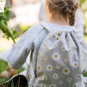 Peut inclure: Un petit sac à dos gris à cordon avec un motif de marguerites blanches. Le sac à dos est fait d'un tissu doux, semblable au lin.
