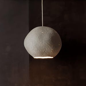 May include: A gray, spherical pendant light with a textured surface. The light fixture is suspended from a thin, white cord. The bottom of the light emits a warm glow, and the surface has small, sparkling details.