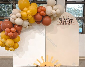 May include: A white and beige backdrop with a balloon arch made of yellow, brown, and cream balloons. The arch is decorated with a wooden sign that says "Here comes the sun THEO TURNS ONE". A wooden sun cutout with the text "@greydesigns.decor" is in the foreground.