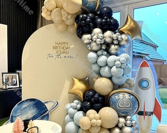 Puede incluir: Una decoración de fiesta de cumpleaños con temática espacial con un arco de globos azules, plateados, dorados y crema. El arco está decorado con planetas y estrellas. El cartel dice "Feliz cumpleaños Jovan, hasta la luna!"