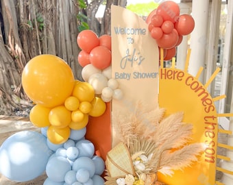 May include: A baby shower decoration featuring a sign that reads "Welcome to Juli's Baby Shower". The decor includes a yellow sun, balloons in shades of yellow, peach, and blue, and a decorative baby carriage.