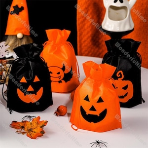 Op de afbeelding: Een verzameling Halloween-taszakken met trekkoord in zwart en oranje. De tassen hebben pompoengezichten en andere griezelige ontwerpen. Een feestelijke kabouter en een spookvormige container staan op de achtergrond.