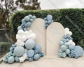 May include: A baby shower backdrop with two beige archways decorated with blue and white balloons. The text "Baby in Bloom" is written on one of the archways.