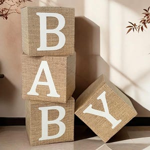 Puede incluir: Cuatro bloques de arpillera beige que deletrean la palabra "BABY" en letras blancas grandes. Los bloques están apilados, con un bloque ligeramente inclinado. El fondo es de color neutro con un toque de luz solar.