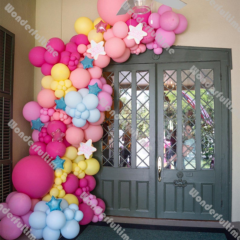 May include: A balloon archway made of pink, yellow, blue, and white balloons. The archway is decorated with star-shaped balloons and is set up in front of a double door.