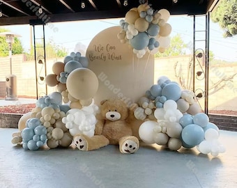 May include: A white backdrop with the text "We Can Bearly Wait" decorated with a garland of blue, white, and beige balloons. A large teddy bear sits in front of the backdrop.