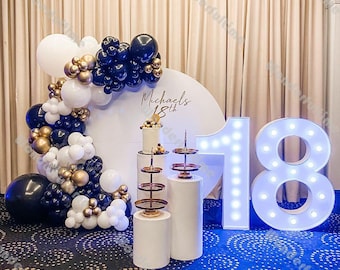 Könnte beinhalten: Weiße, schwarze und goldene Ballons, die zu einer Girlande um einen weißen Hintergrund mit dem Text "Michaels 18th" angeordnet sind. Zwei weiße Podest mit mehrstöckigen Kuchenständern und einer weißen leuchtenden Nummer 18.