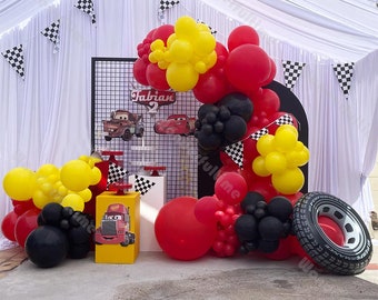 May include: A Cars-themed birthday party decoration with red, yellow, and black balloons, a black metal grid backdrop with a sign that says "Fabian 2", and a yellow cake stand with a red and yellow toy truck.