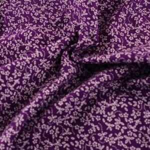 May include: Close-up of a purple fabric with a repeating pattern of small, white floral designs. The fabric is gathered, showing folds and shadows that highlight the texture and detail of the print. The floral pattern is evenly distributed across the material.